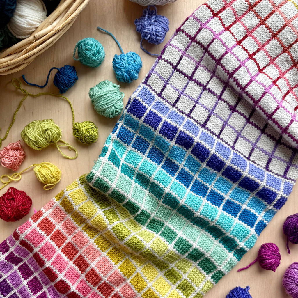 A hand holds a black box of the Pacific Knit Co. Geometric Doodle Card Deck, surrounded by colorful yarn balls and a plaid blanket.