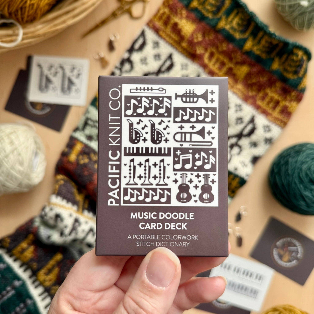 A hand holds a small box labeled 'PACIFIC KNIT CO. MUSIC DOODLE CARD DECK' against a styled flat-lay background of knitting yarn, cards, and a patterned textile.