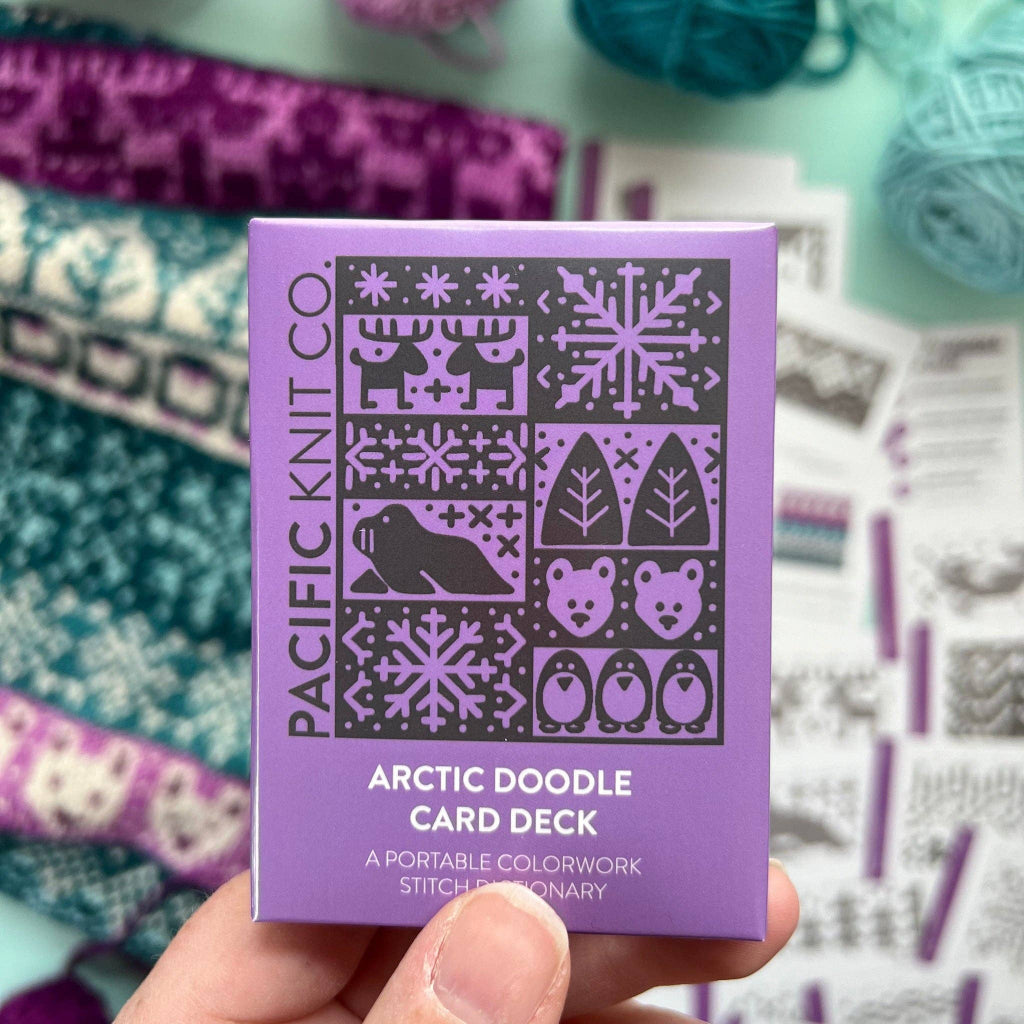A hand holding a purple box of Pacific Knit Co. Arctic Doodle Card Deck, with knitting yarn and pattern sheets blurred in the background.