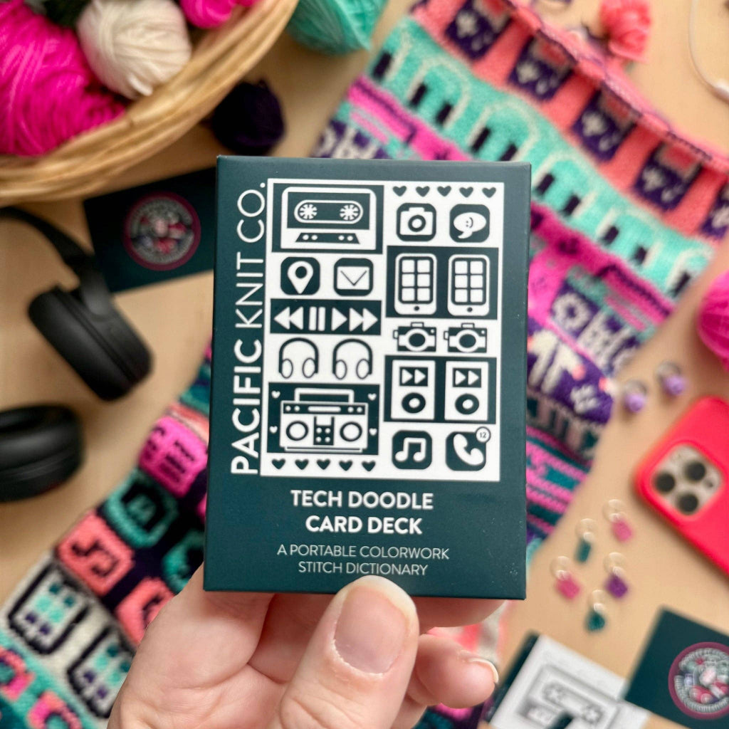 A hand holds a dark teal box labeled 'PACIFIC KNIT CO. TECH DOODLE CARD DECK' against a cozy, colorful background of knitted fabrics, yarn, and accessories.
