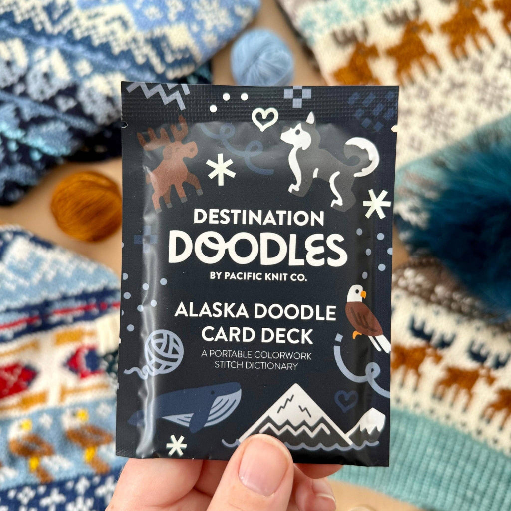 A hand holds a small product card for the Destination Doodles Alaska Doodle Card Deck by Pacific Knit Co., with a cozy, patterned knit blanket in the background.