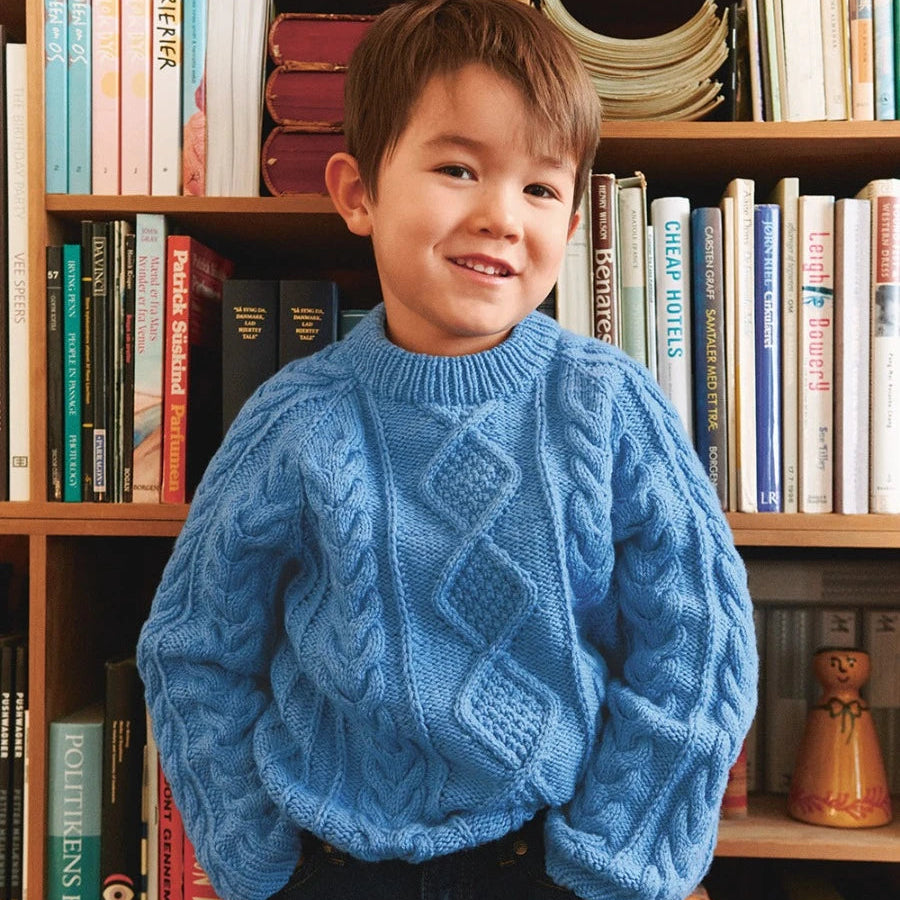 Boy wearing a blue cable sweater standing in front of a bookshelf filled with books.