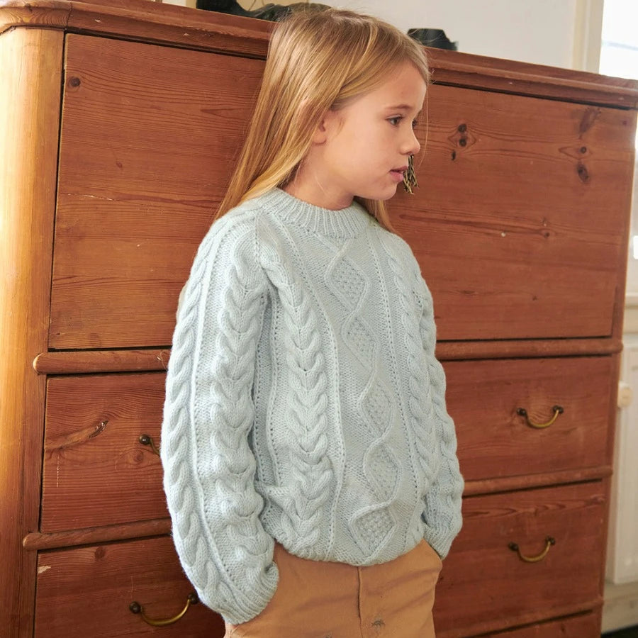 Young girl wearing a light blue cable knit sweater standing in front of a wooden dresser.