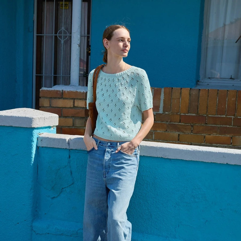 Woman in light blue patterned top and jeans standing against a blue wall.