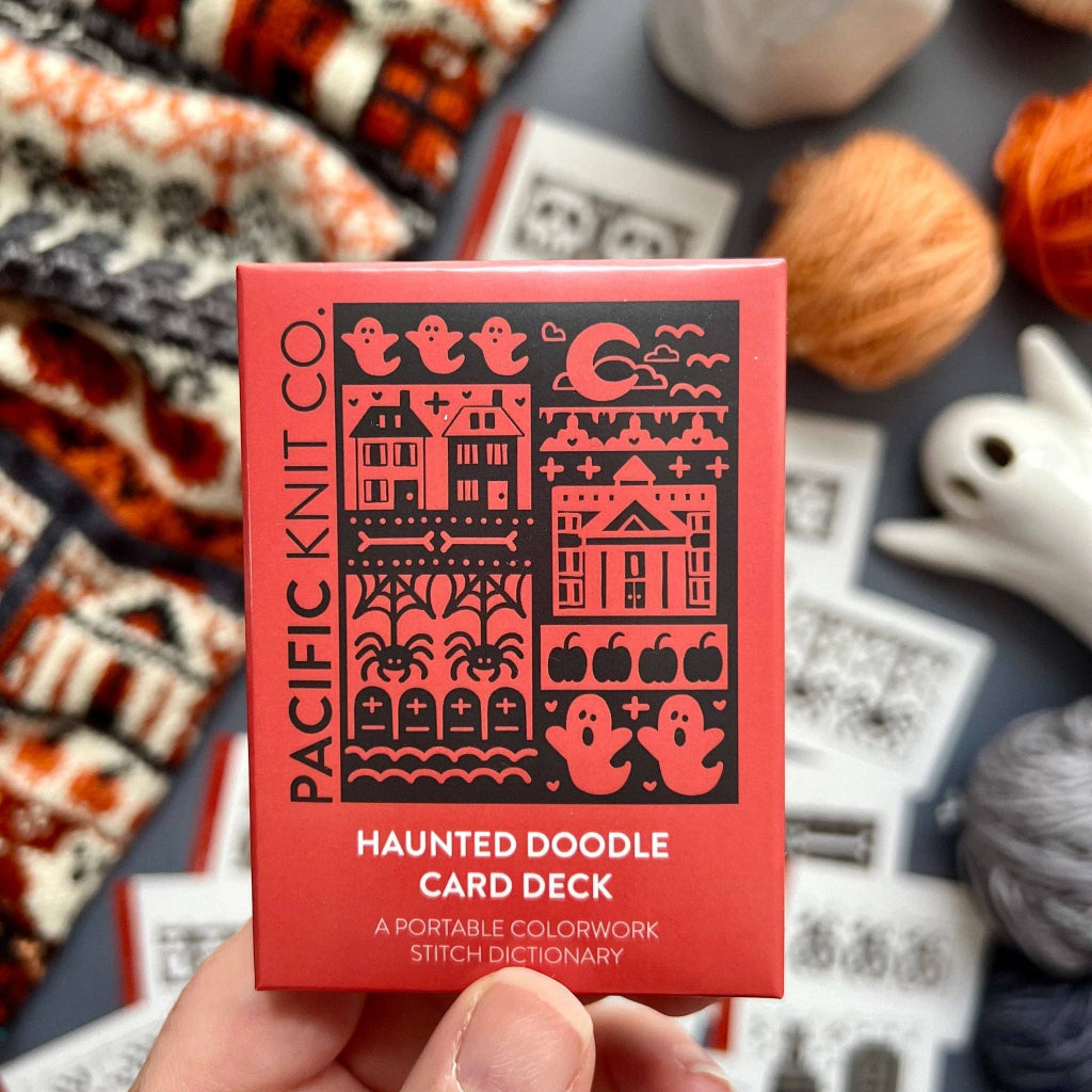A hand holds a red box of the Pacific Knit Co. Haunted Doodle Card Deck, surrounded by knitting supplies like yarn balls and stitch cards on a gray surface.