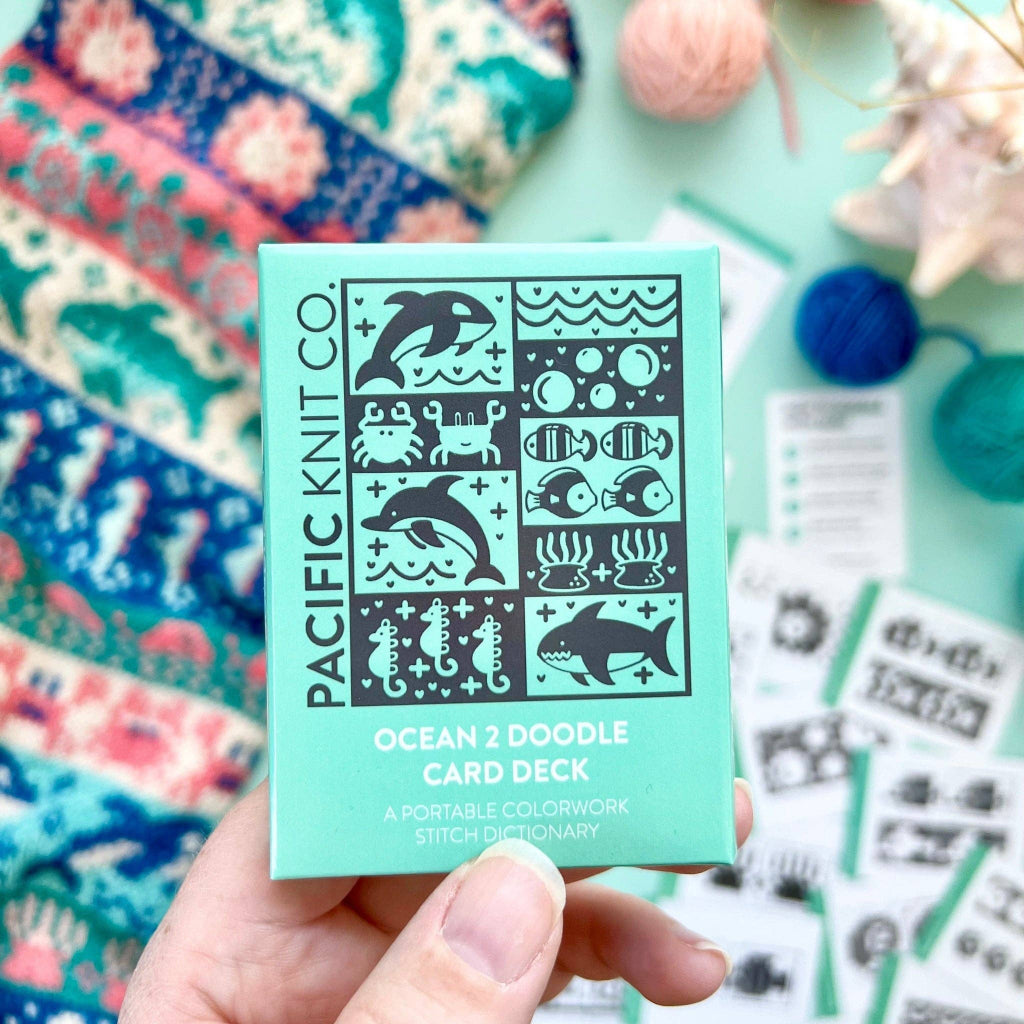 A hand holds a mint green box labeled 'PACIFIC KNIT CO. OCEAN 2 DOODLE CARD DECK' against a styled flat-lay background with colorful yarn balls, fabric, and sewing supplies.