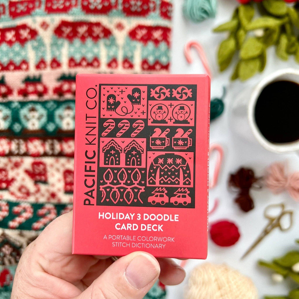 A hand holds a pink and black box of Pacific Knit Co. Holiday 3 Doodle Card Deck against a cozy, blurred background of knitted sweaters, yarn, and a cup of coffee.