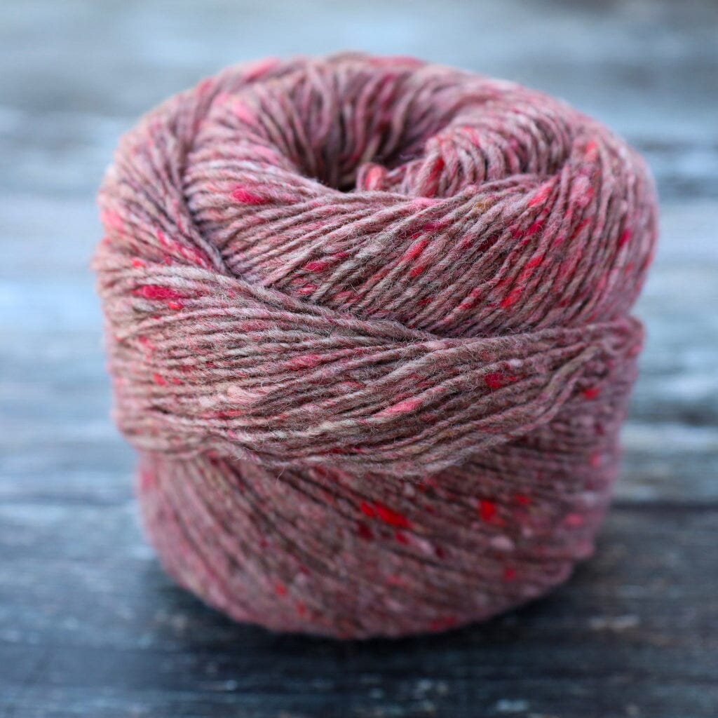 A ball of heathered dusty rose and brown yarn with small red flecks, photographed on a dark rustic wooden surface.
