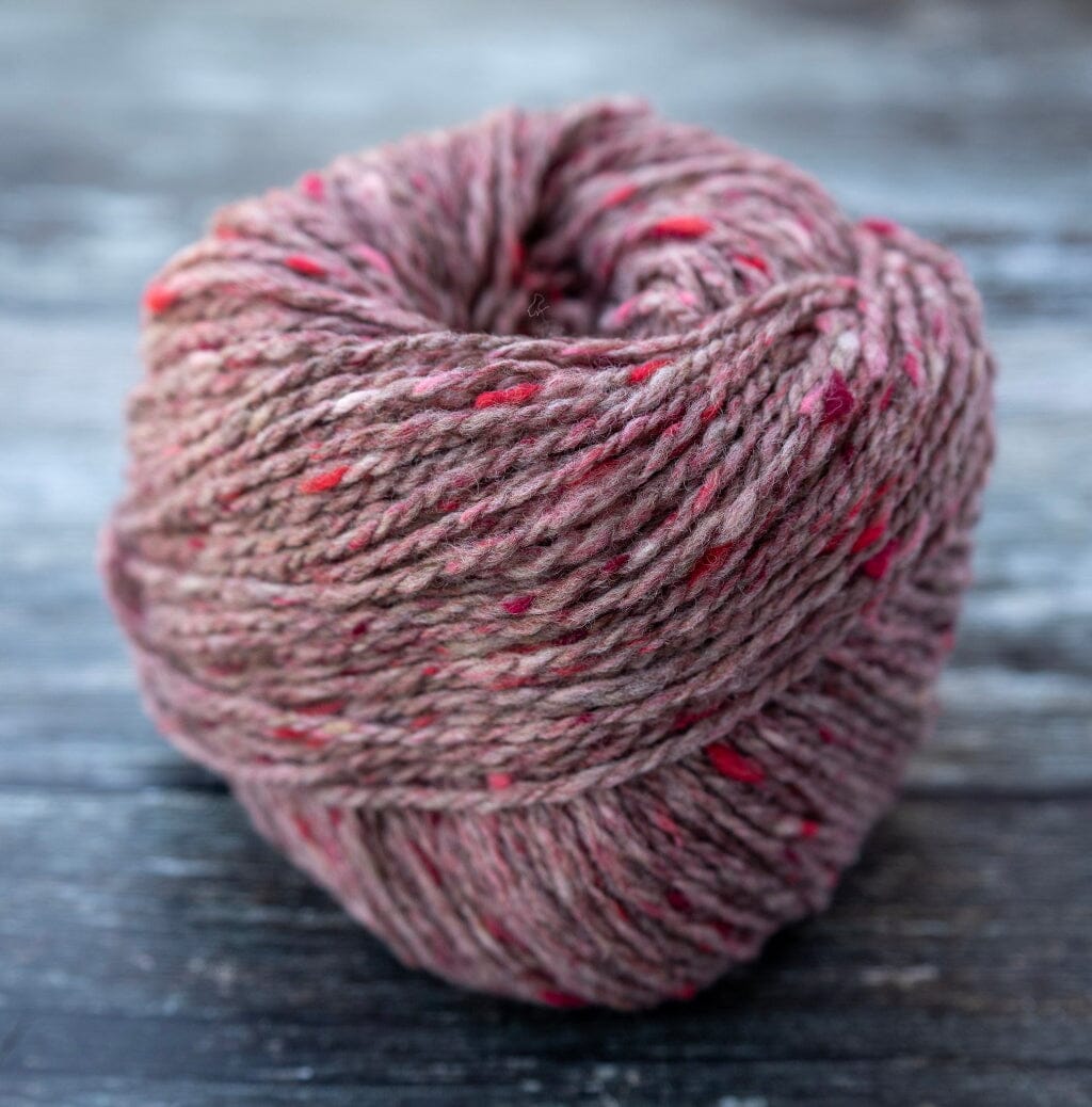 A ball of variegated yarn in a dusty mauve-brown shade with scattered bright pink and red flecks, photographed on a dark weathered wooden surface.