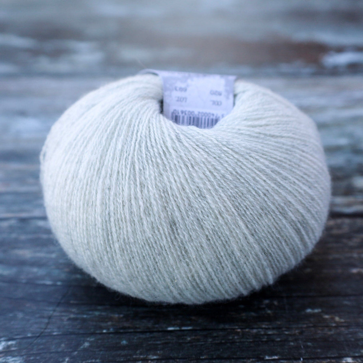 A single ball of light gray yarn resting on a dark, weathered wooden surface. A paper label is visible at the top of the ball.