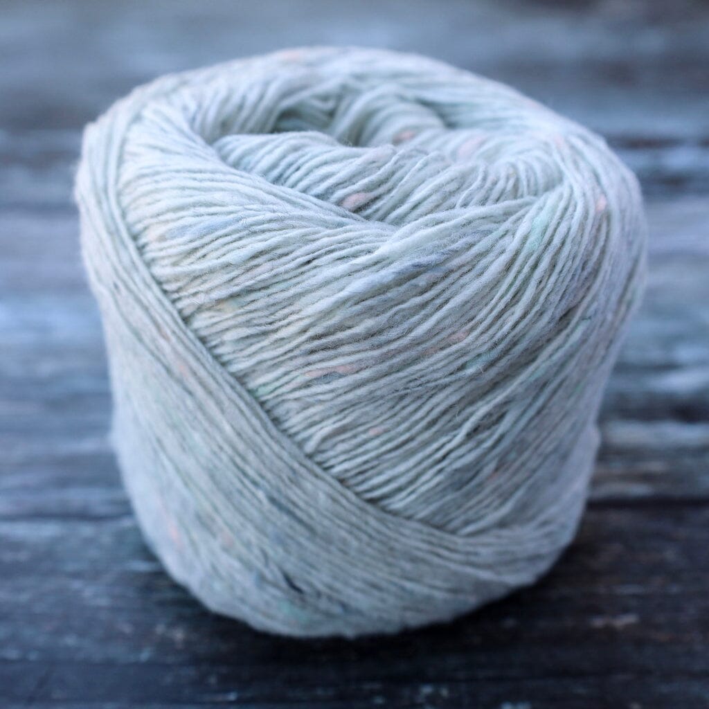 A ball of variegated yarn in a muted sage green-gray color with subtle pastel flecks, resting on a dark rustic wooden surface.