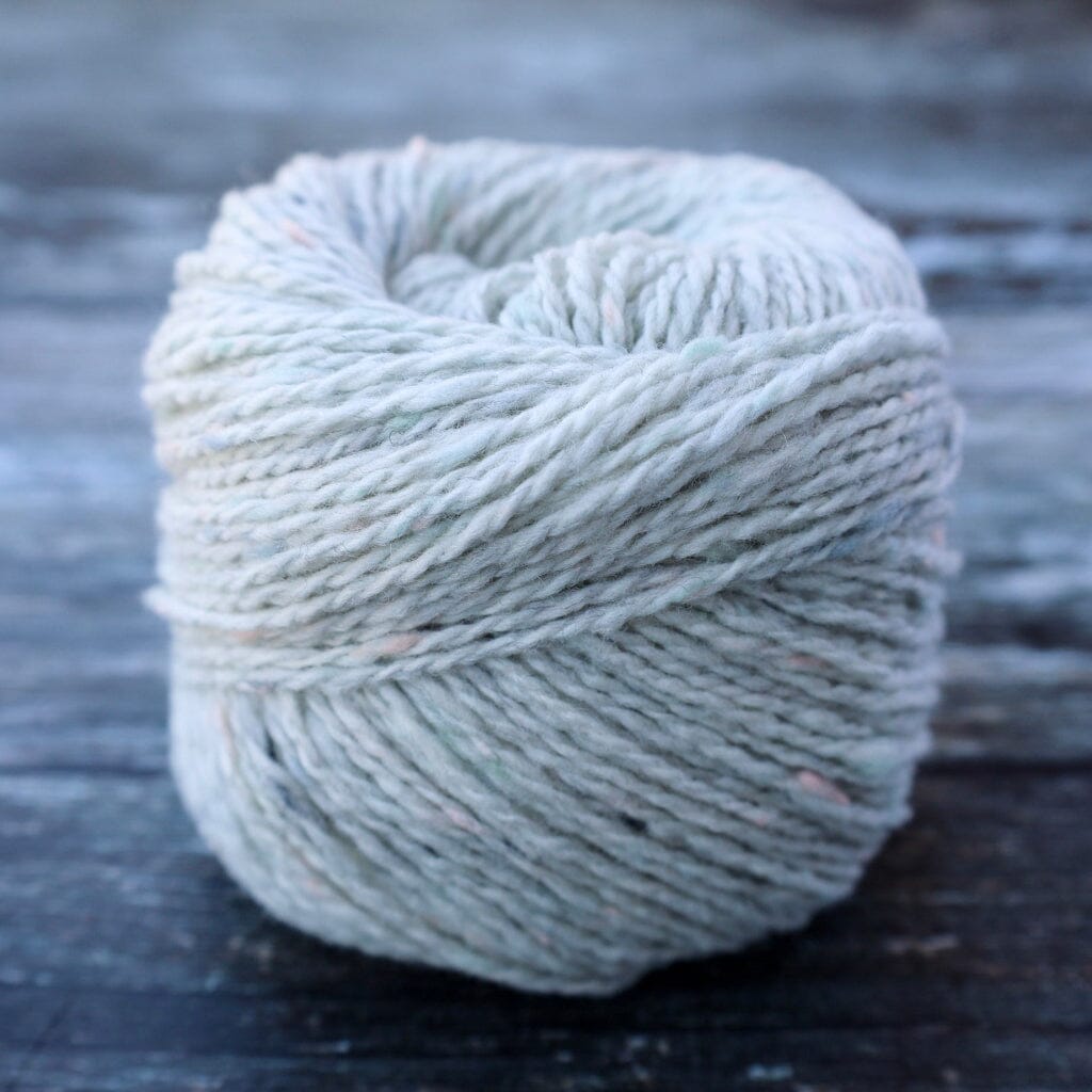A ball of variegated yarn in a muted sage green and pale blue-gray colorway, resting on a dark, weathered wooden surface.