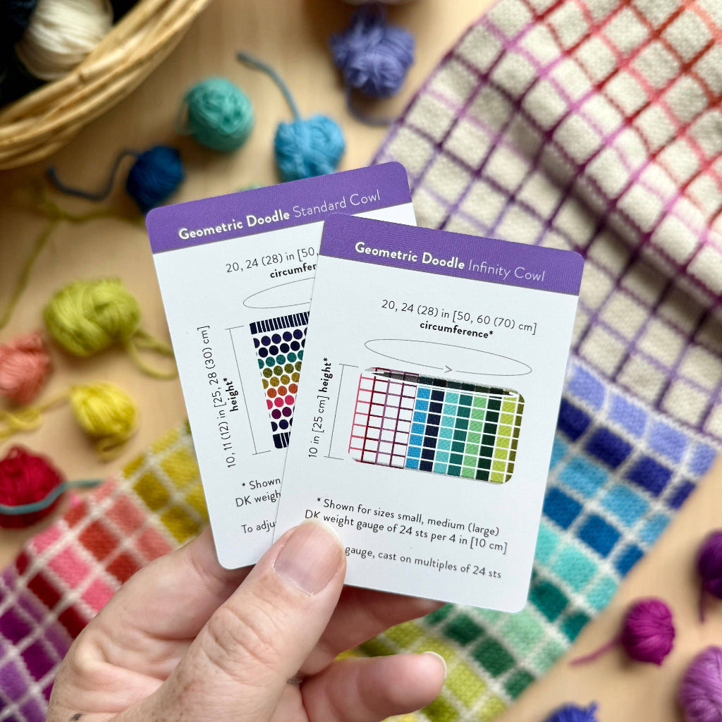 A hand holds two small printed cards with purple headers and colorful geometric charts, photographed over a plaid blanket with yarn balls and a basket in the background.