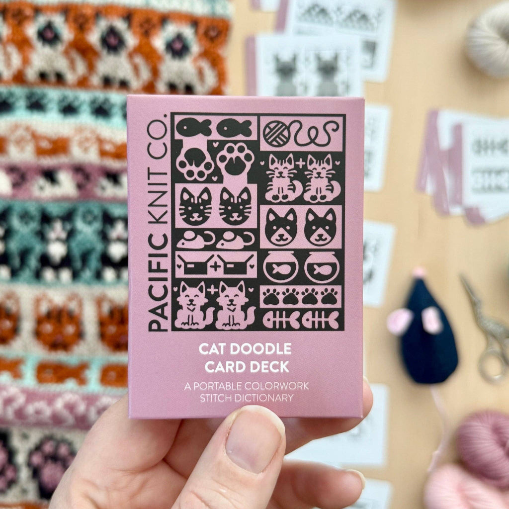 A hand holds a pink box labeled 'PACIFIC KNIT CO. CAT DOODLE CARD DECK' against a softly blurred craft table background with yarn and sewing supplies.