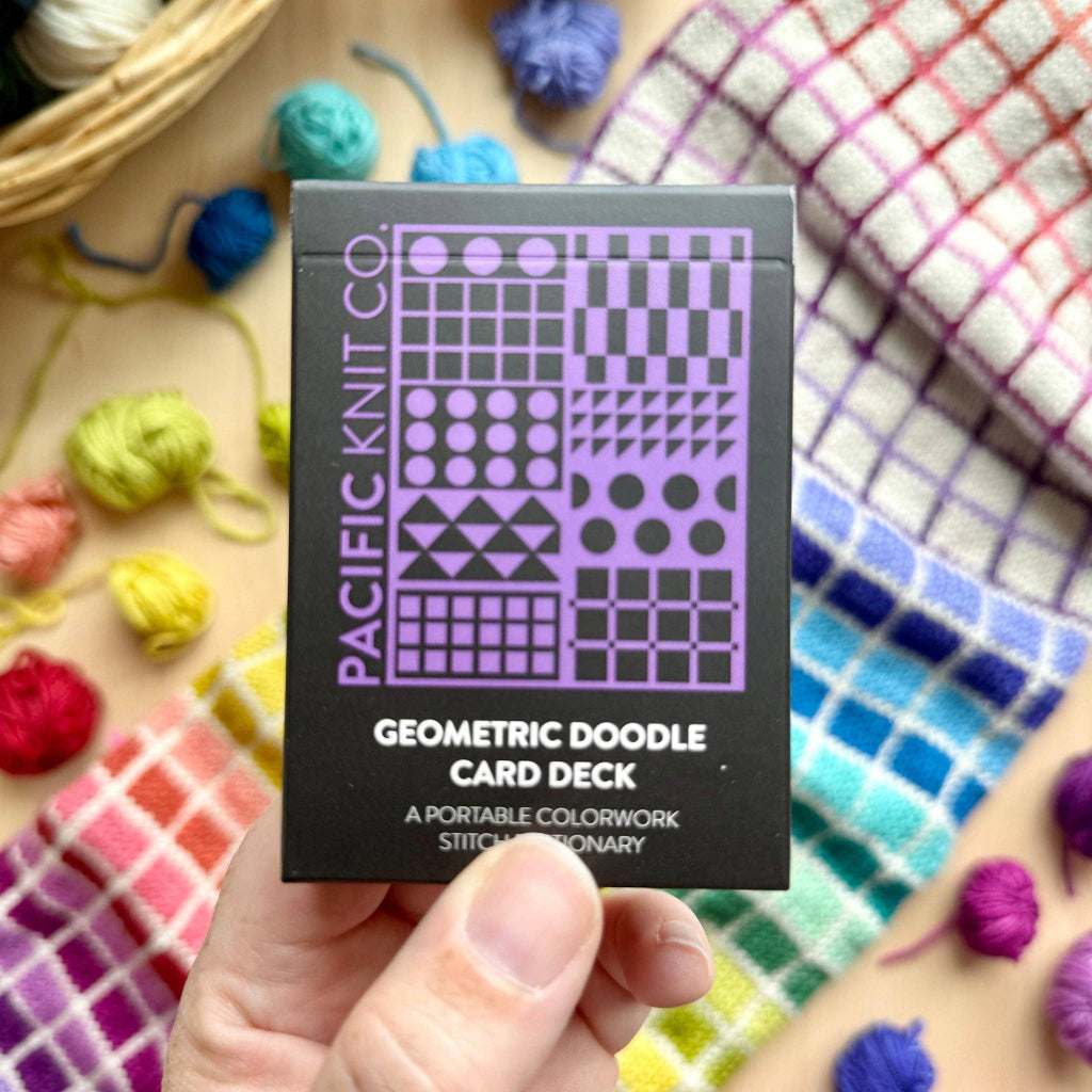 A hand holds a black box of the Pacific Knit Co. Geometric Doodle Card Deck, surrounded by colorful yarn balls and a plaid blanket.