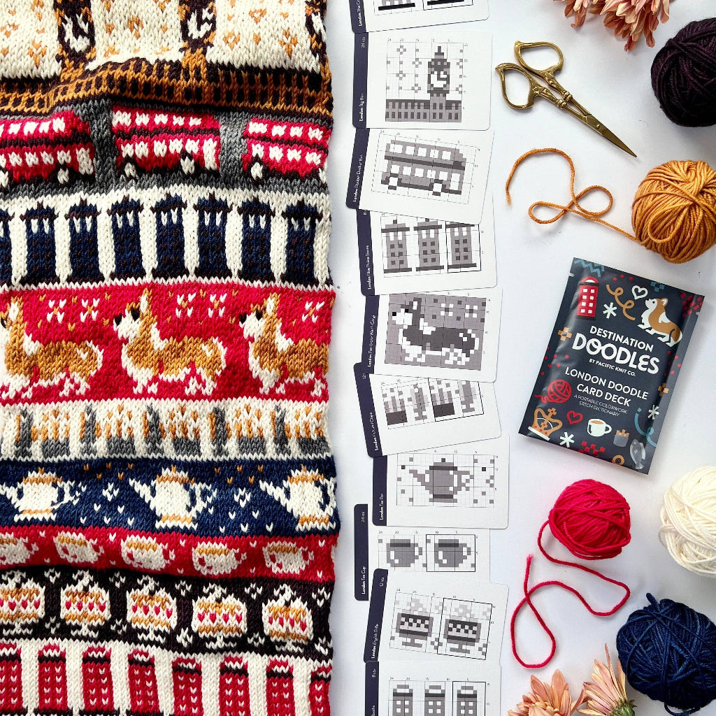 A flat-lay of a knitted scarf with colorful London-themed patterns, alongside printed pattern cards, yarn balls, scissors, and flowers on a white surface.