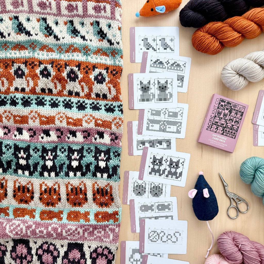 A hand holds a pink box labeled 'PACIFIC KNIT CO. CAT DOODLE CARD DECK' against a softly blurred craft table background with yarn and sewing supplies.