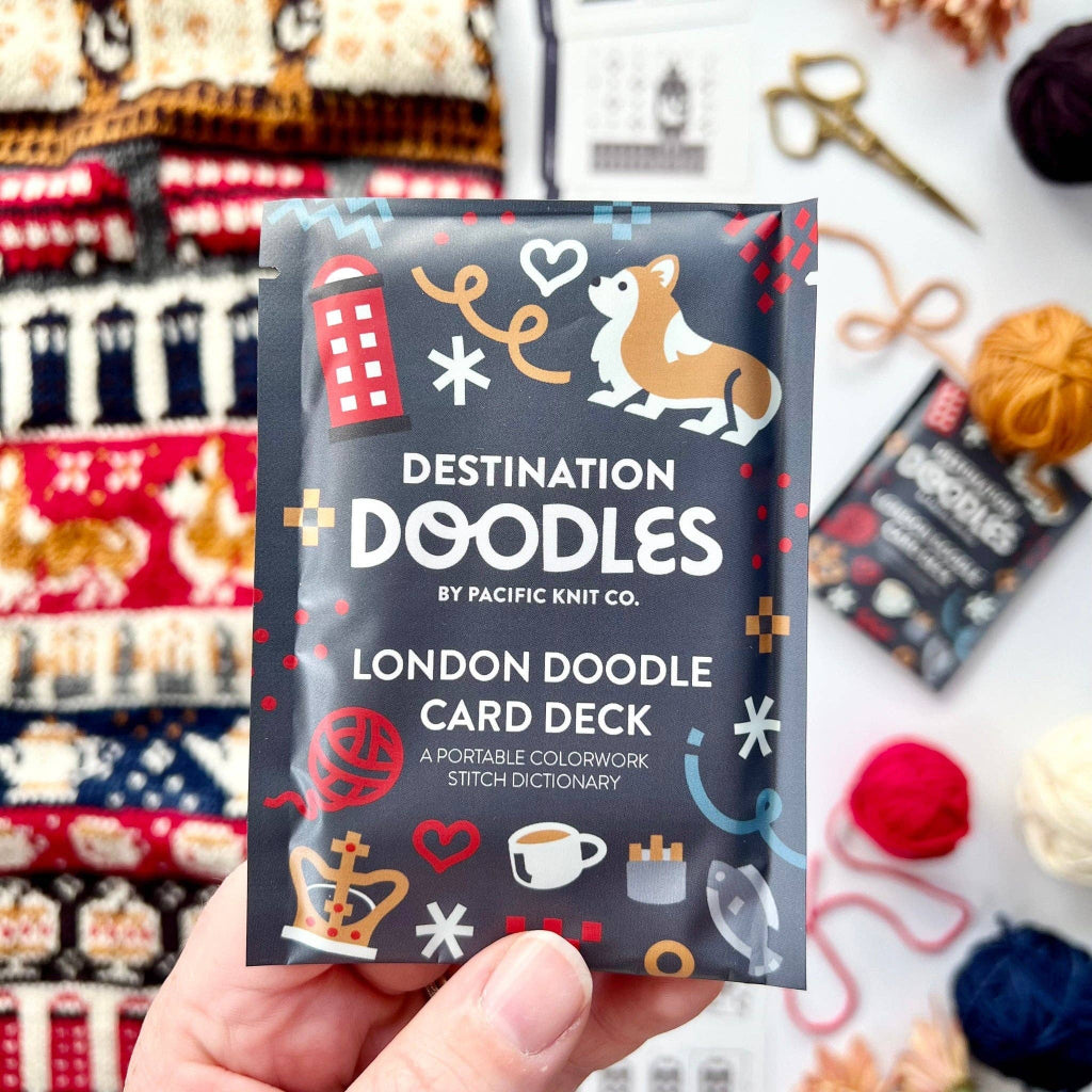 A hand holds a 'Destination Doodles' London Doodle Card Deck packet in front of a white background decorated with knitting needles, yarn balls, and knitted garments. The packet is dark gray with colorful doodles of a corgi, a red telephone booth, and other London-themed illustrations.