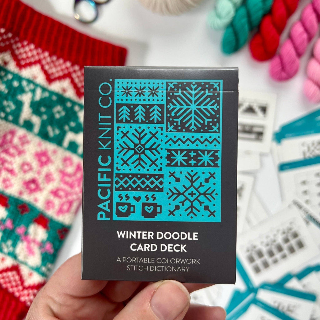 A hand holds a black box of Pacific Knit Co. Winter Doodle Card Deck, featuring a teal and black winter pattern graphic on the front.