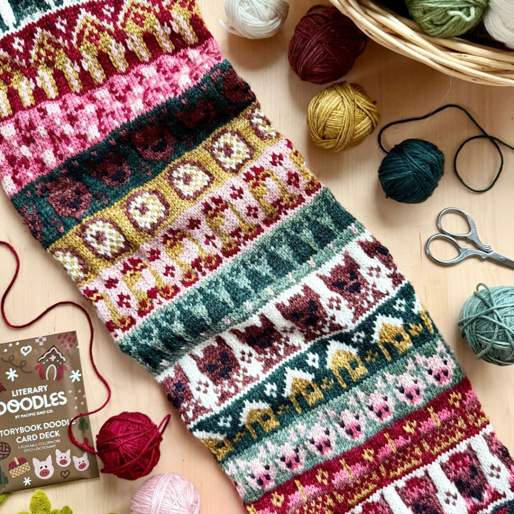 A close-up of a knitted scarf with a colorful Fair Isle pattern featuring snowflakes, trees, and geometric motifs in pink, green, red, yellow, and white. The scarf is laid out on a light wood surface surrounded by knitting supplies, including balls of yarn, scissors, and a card deck box labeled 'Literary Doodles'.