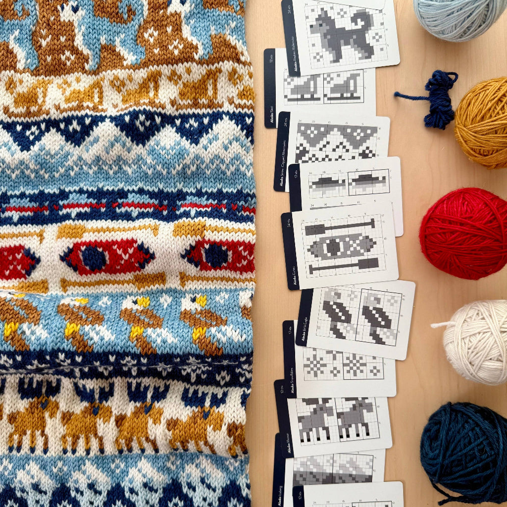 A knitted textile with repeating geometric and animal motifs in blue, cream, brown, red, and yellow is shown beside a row of pattern cards on a light wood surface, with balls of yarn around them.