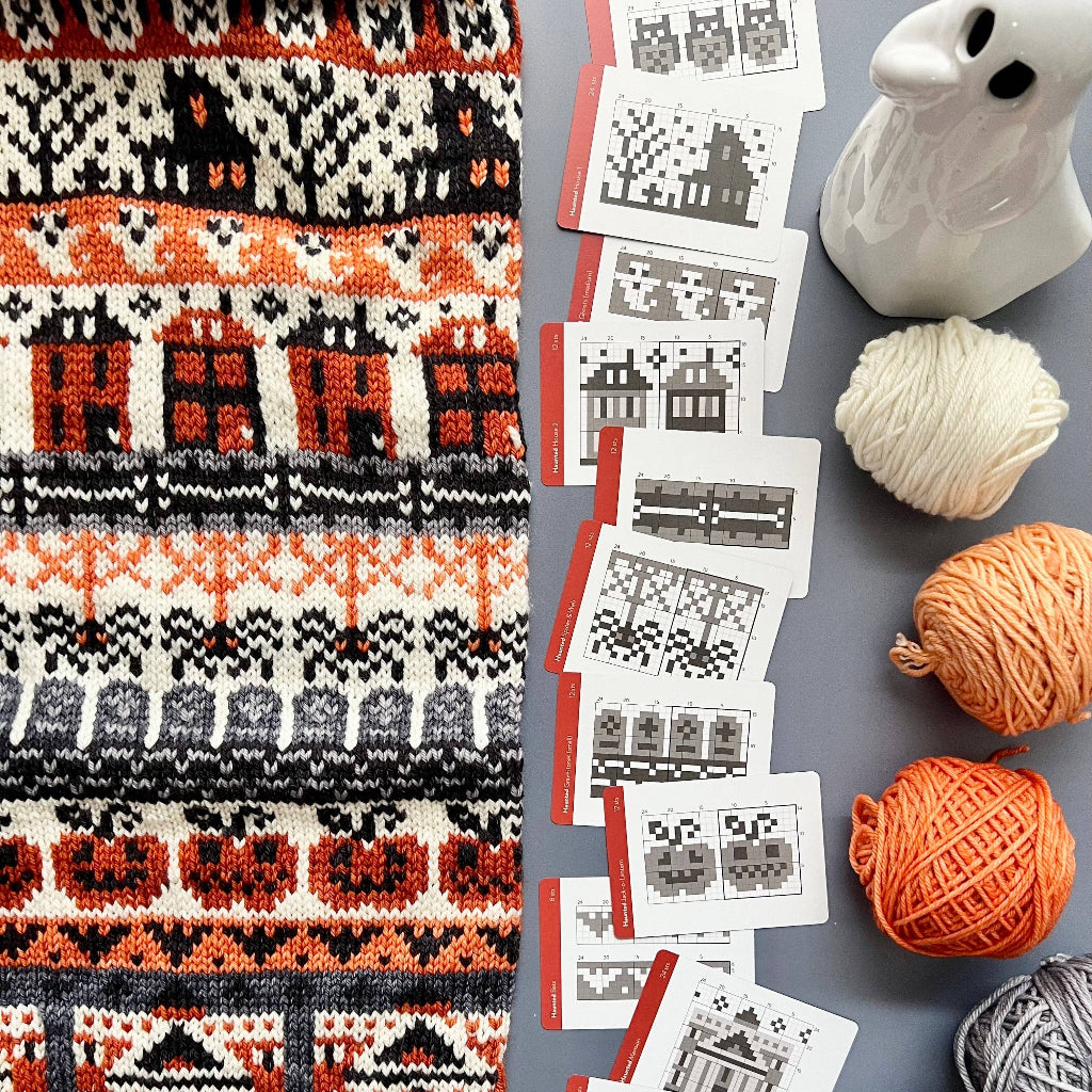 A flat-lay of a knitted textile with a repeating house pattern in orange, black, and cream, arranged alongside yarn balls, printed pattern cards, and a white ceramic ghost figurine on a gray surface.