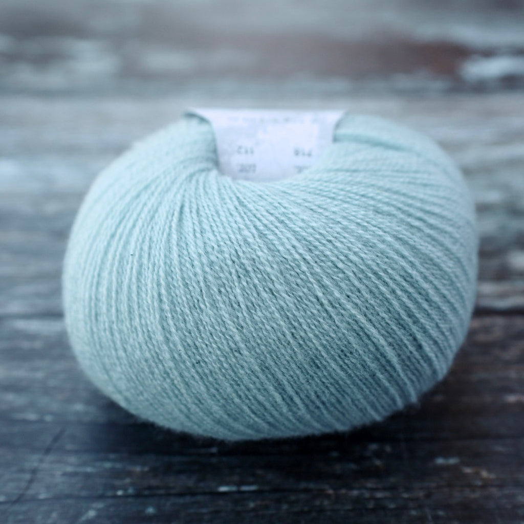 A ball of light blue-gray yarn resting on a dark rustic wooden surface, with a small white label visible at the top.