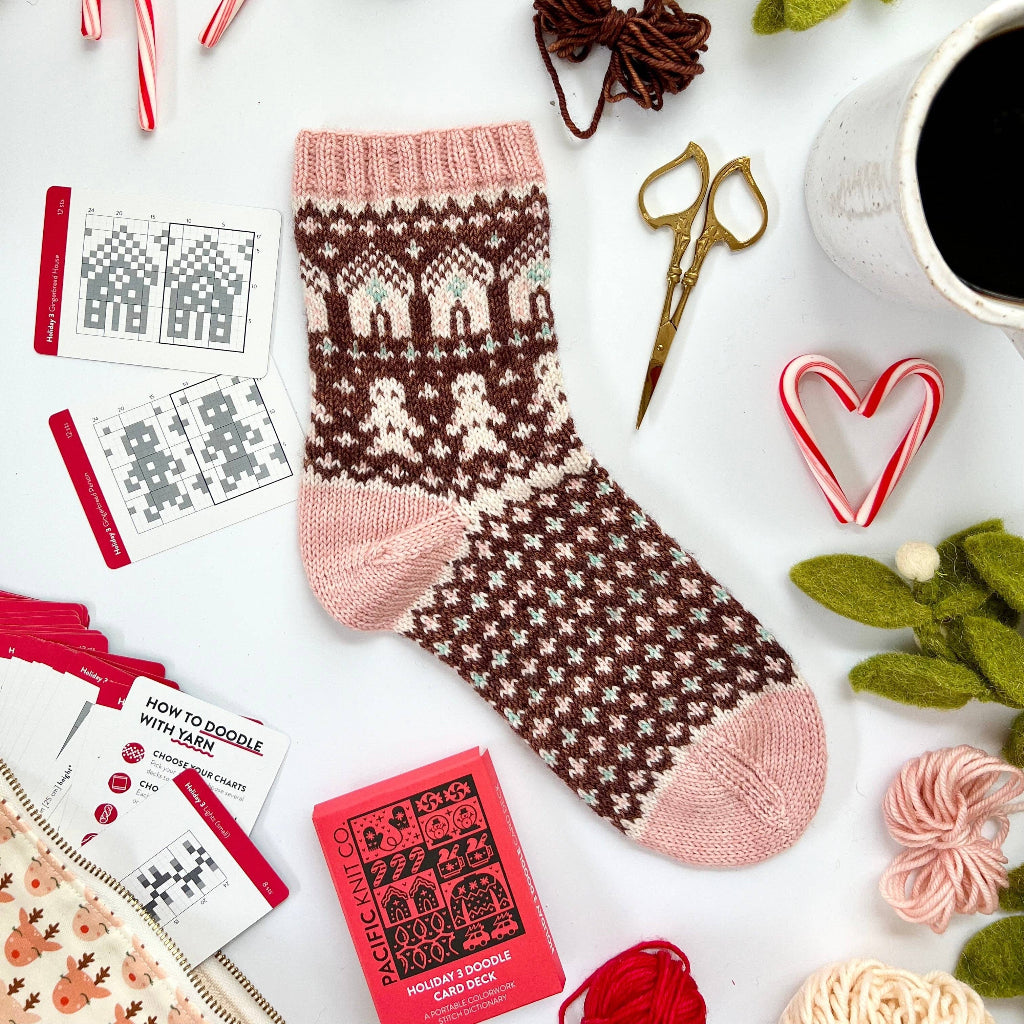 A festive Christmas sock with a brown and pink Fair Isle pattern is displayed on a white background surrounded by knitting supplies, candy canes