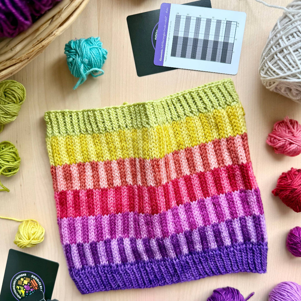 A handmade knitted cowl with horizontal rainbow stripes in yellow, green, pink, red, purple, and blue, displayed on a light wood background surrounded by yarn balls and knitting cards.