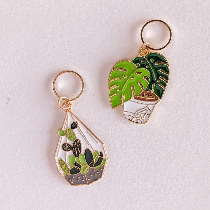 Two gold-tone enamel plant keychains on a light off-white background. One keychain features a teardrop-shaped terrarium design with green cacti and succulents, and the other features a potted Monstera plant with a green leaf and white pot.