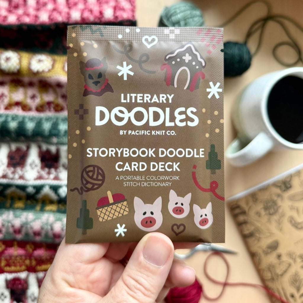 A hand holds a packet of Literary Doodles Storybook Doodle Card Deck by Pacific Knit Co. The brown packet features cute illustrations of pigs, a house, and knitting motifs, set against a cozy background of knitted fabrics, a cup of coffee, and yarn.