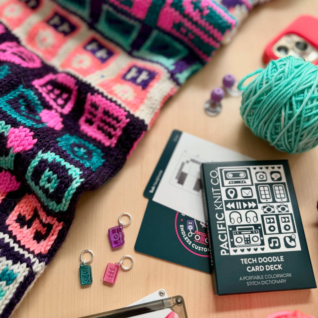Colorful knitted items and yarn on a wooden surface with a card deck and small charms.