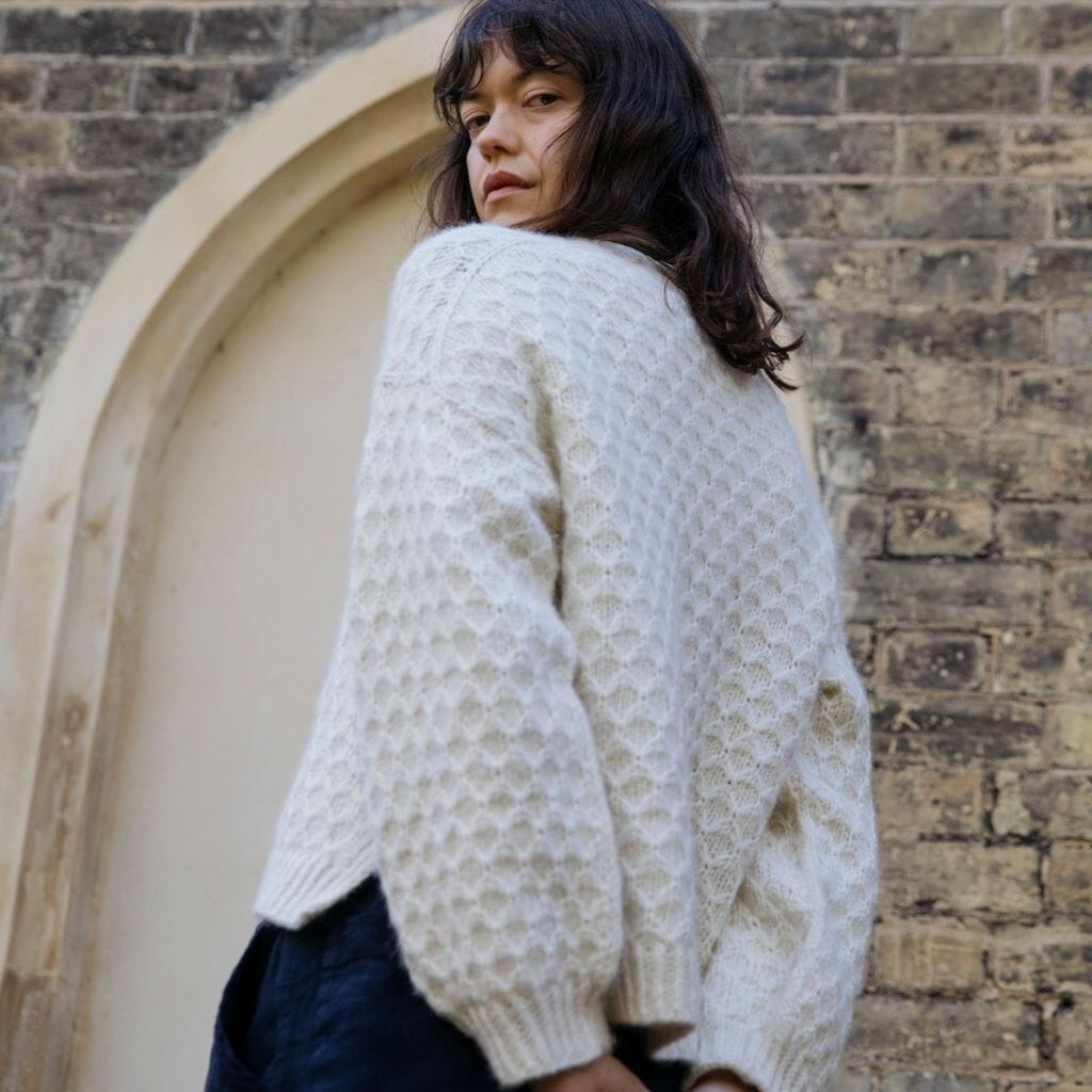 Person wearing a patterned knitted sweater standing against a brick wall.