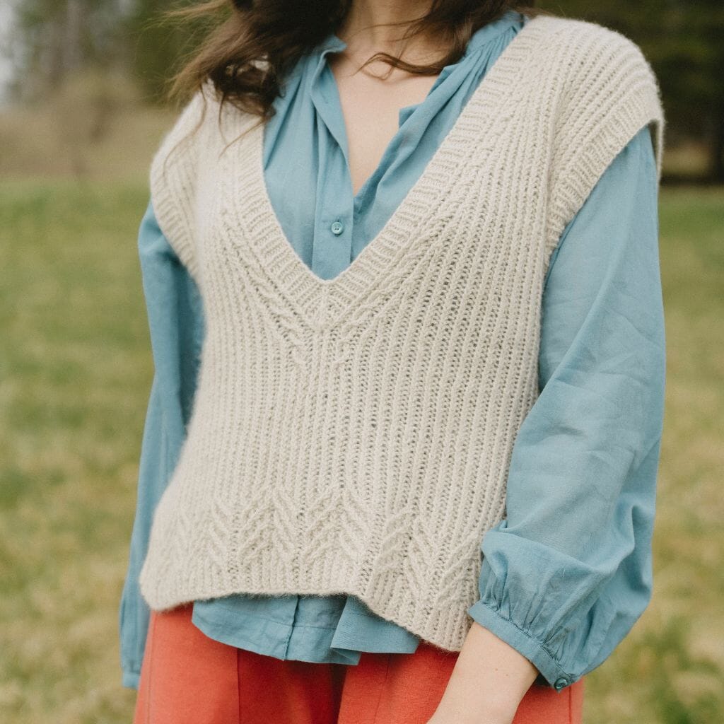Person wearing a beige knitted vest over a blue shirt with a blurred natural background