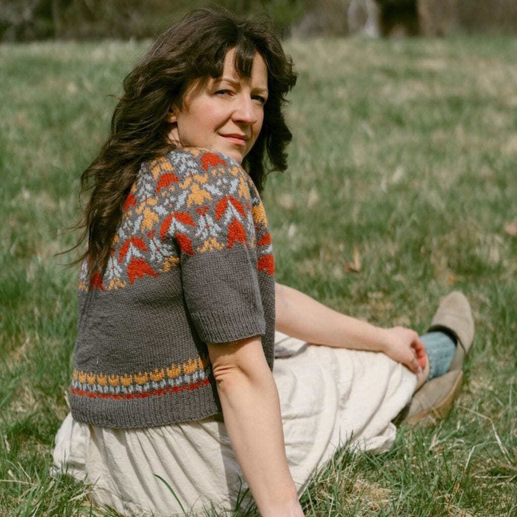 Woman sitting in a grassy field wearing a colourwork knitted top.