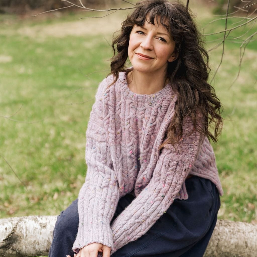 Woman wearing a pink knitted cable sweater sitting on a log in a park.