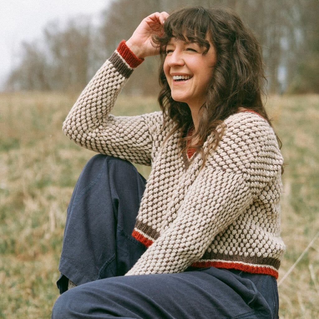 Woman wearing a chunky knit sweater sitting outdoors on a grassy hill.