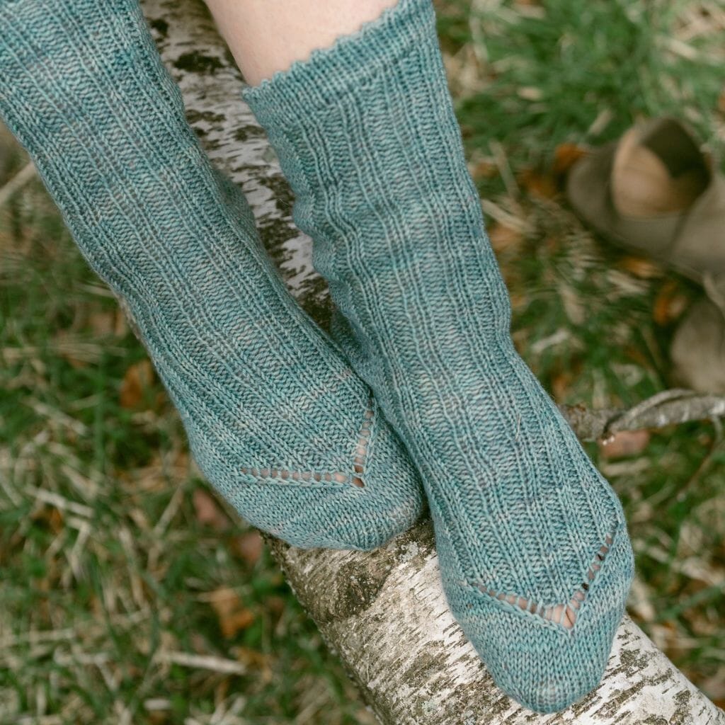 Teal knitted socks worn by a person sitting on a tree branch with grass and leaves in the background.