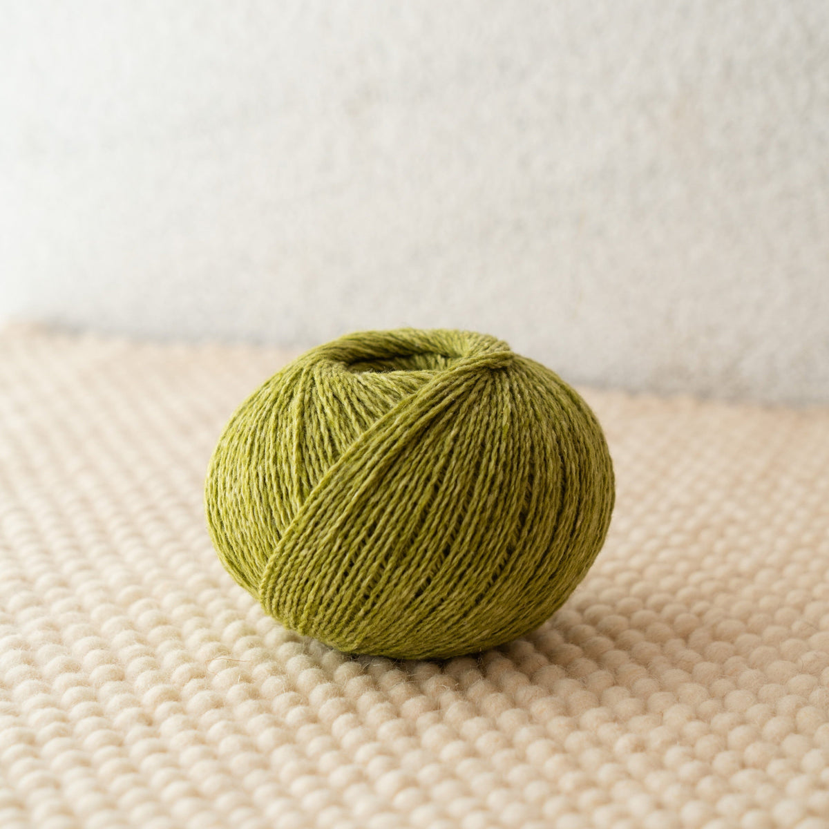 A ball of olive green yarn resting on a textured cream woven surface against a soft off-white background.