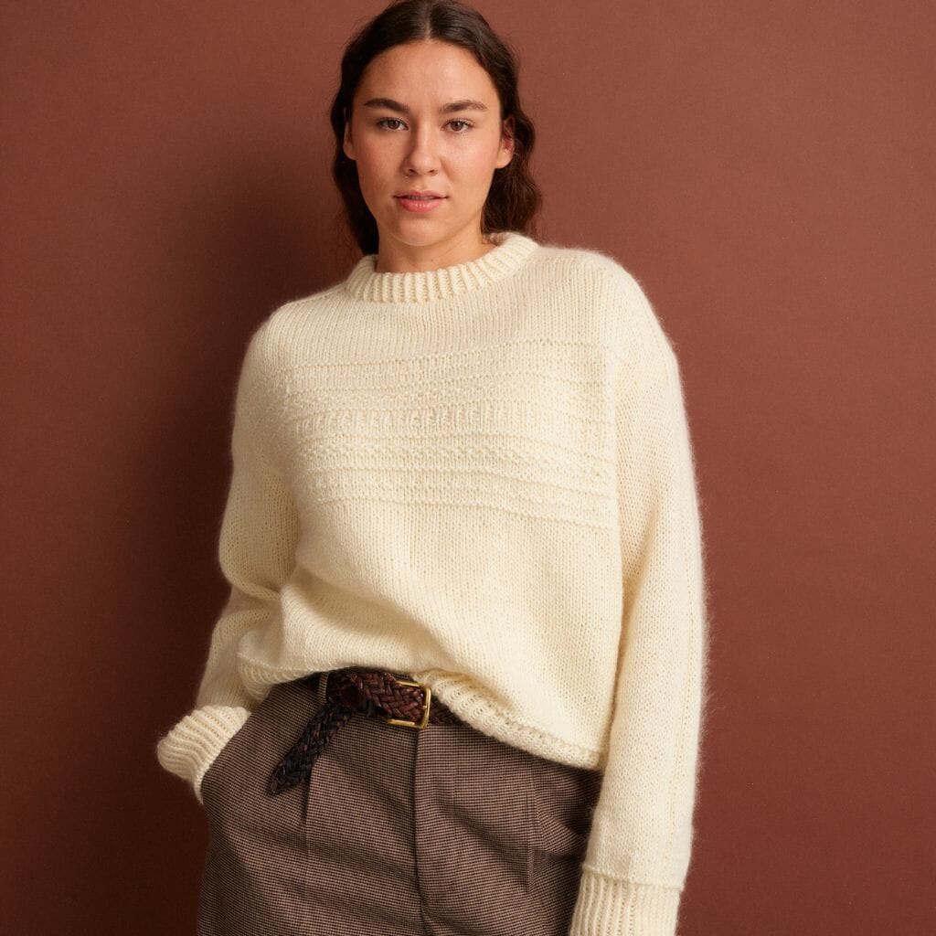 Woman wearing a cream sweater and brown pants against a brown background