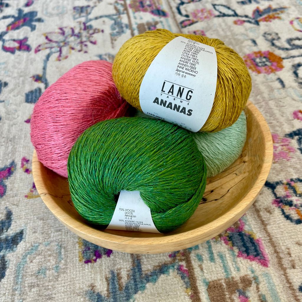 A bowl containing four balls of Lang Yarns Ananas yarn in pink, green, and yellow colors, placed on a patterned surface.