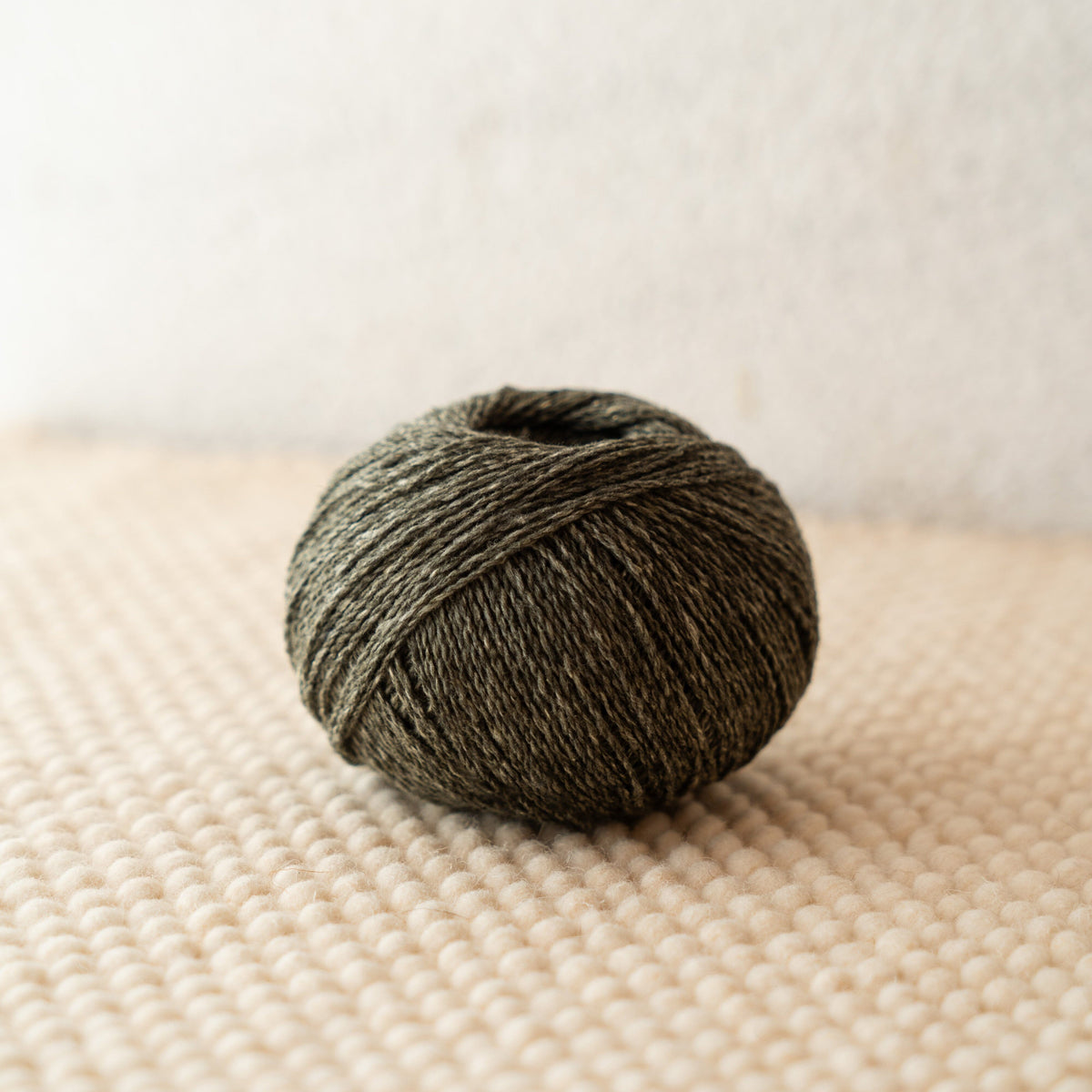 A ball of dark olive green yarn resting on a textured beige woven surface against a soft off-white background.