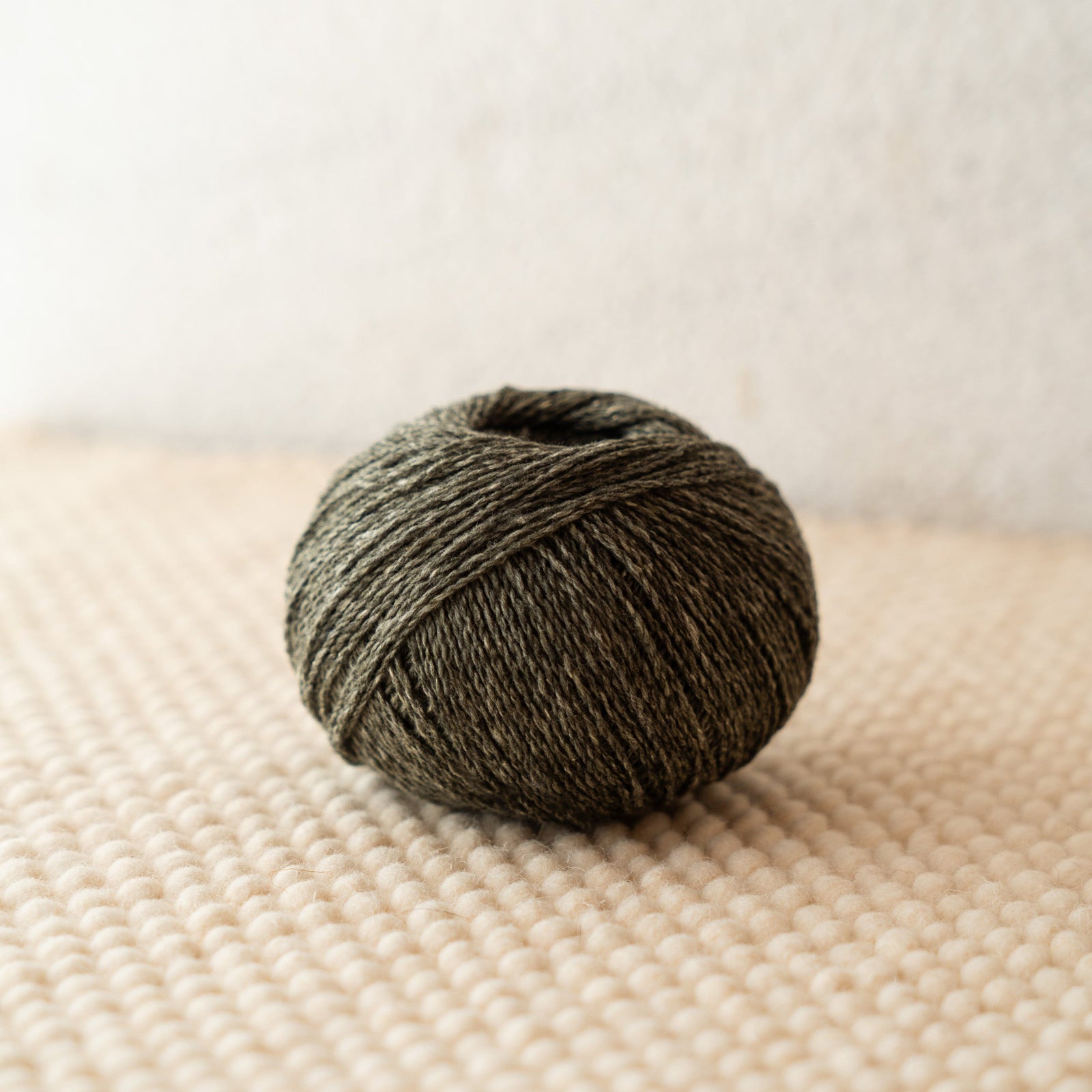 A ball of dark olive green yarn resting on a textured beige woven surface against a soft off-white background.