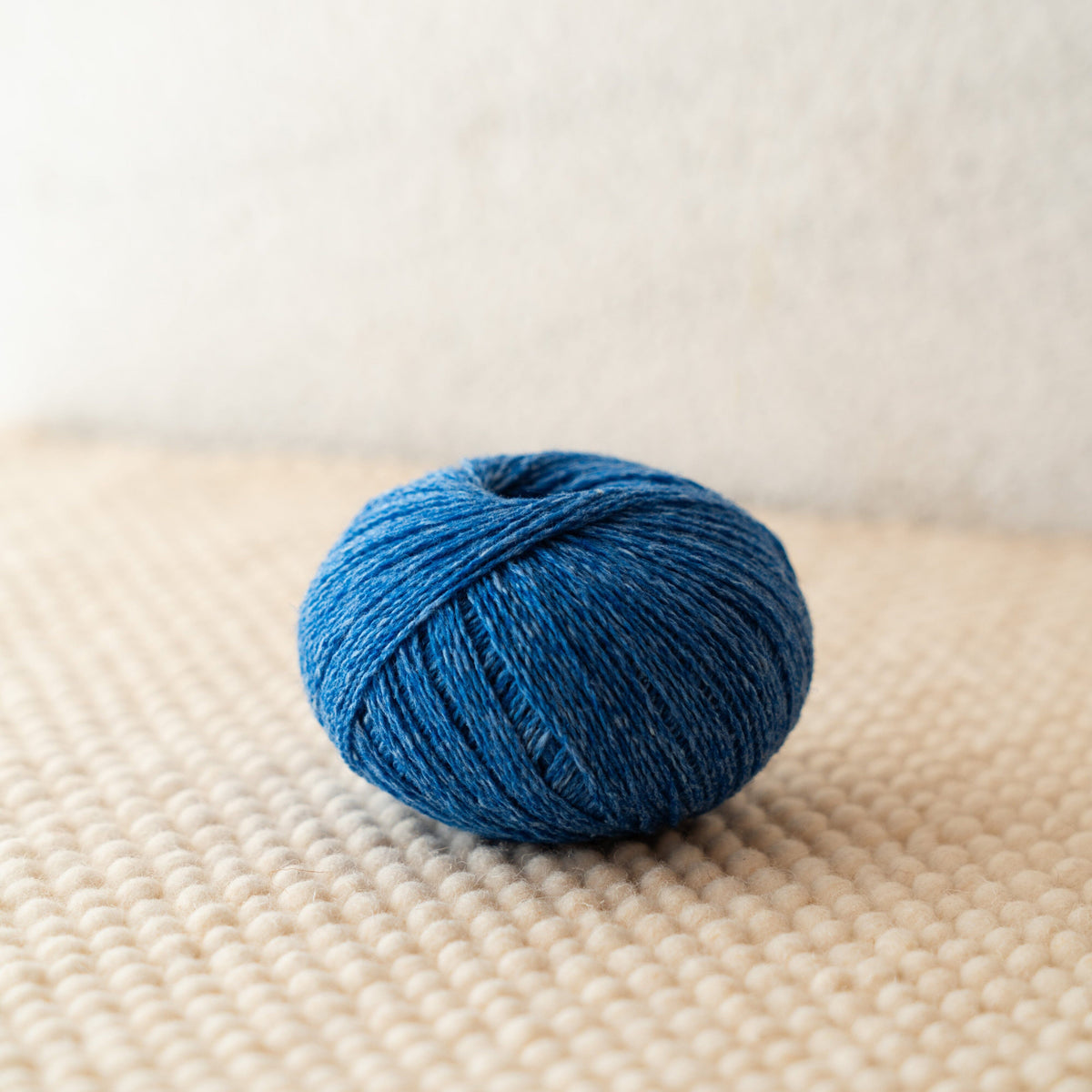 A small ball of textured blue yarn resting on a beige woven surface against a soft off-white background.