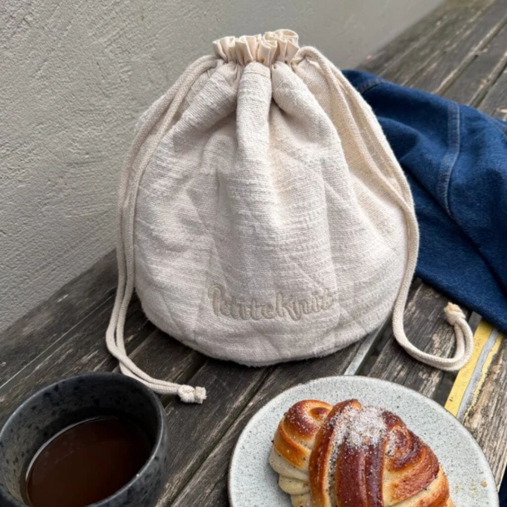 A cream-colored cotton drawstring pouch with 'PetiteKnit' embroidered on the front, styled on a rustic wooden table next to a cup of coffee, a pastry on a plate, and a blue denim jacket.