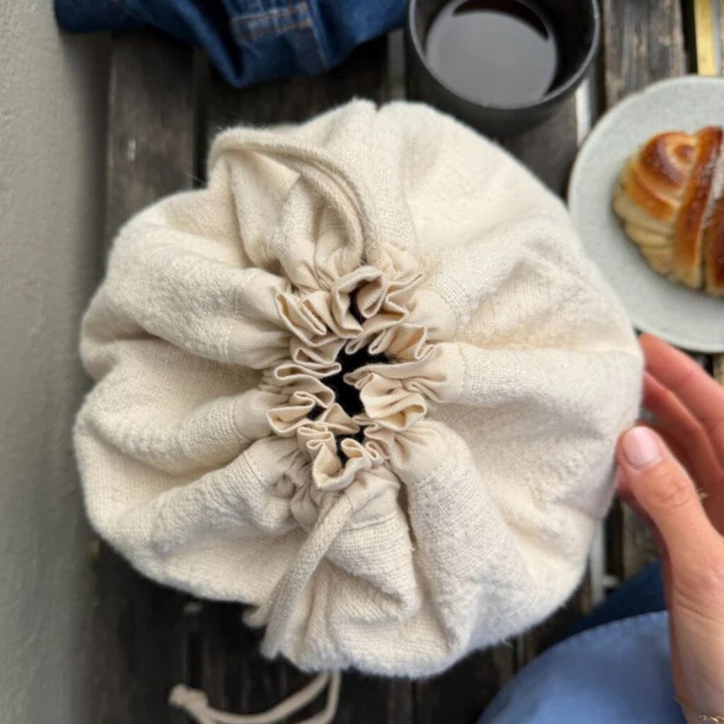 A cream-colored cotton drawstring bag held by a hand, set on a rustic wooden table next to a cup of coffee and a pastry.