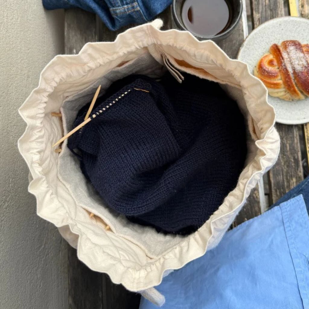 An off-white ruffled drawstring bag filled with a navy blue knit sweater, shown from a top-down angle on a rustic wooden table with a pastry and coffee nearby.