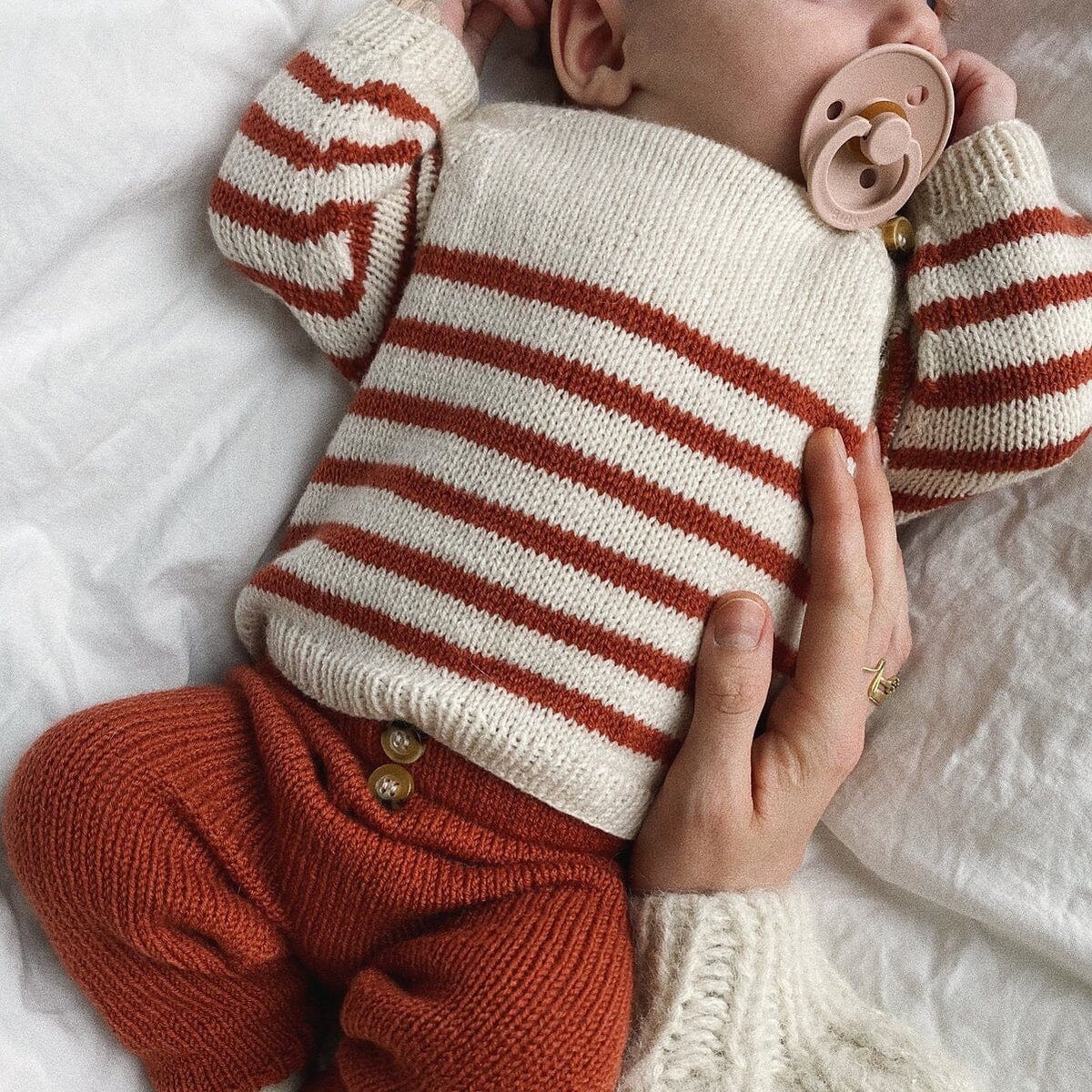 Baby in striped sweater and rust-colored pants being held on a white blanket
