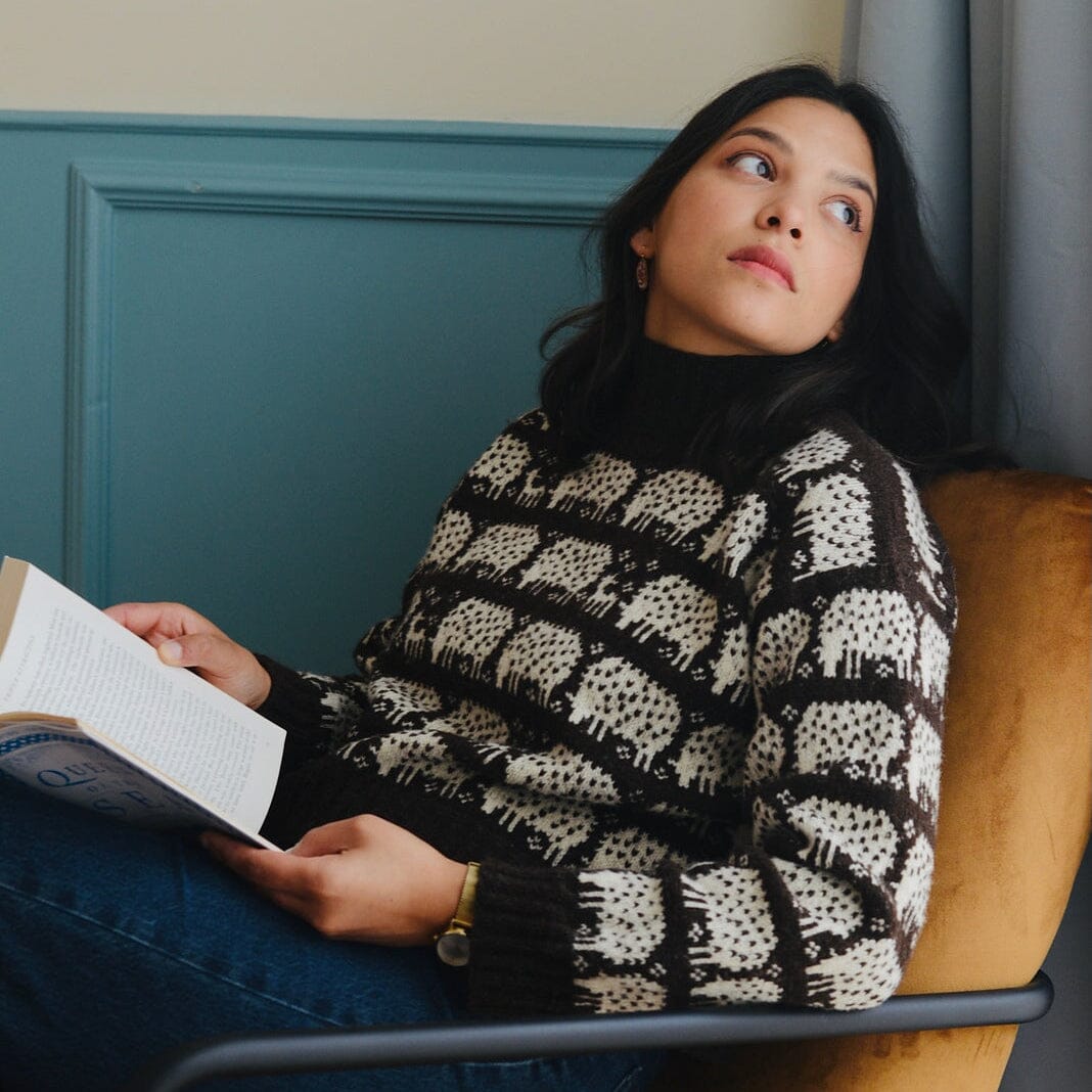 A woman sits in a mustard-yellow armchair, looking upward while holding an open book. She is wearing a black and cream patterned knit sweater over a black turtleneck, paired with blue jeans.