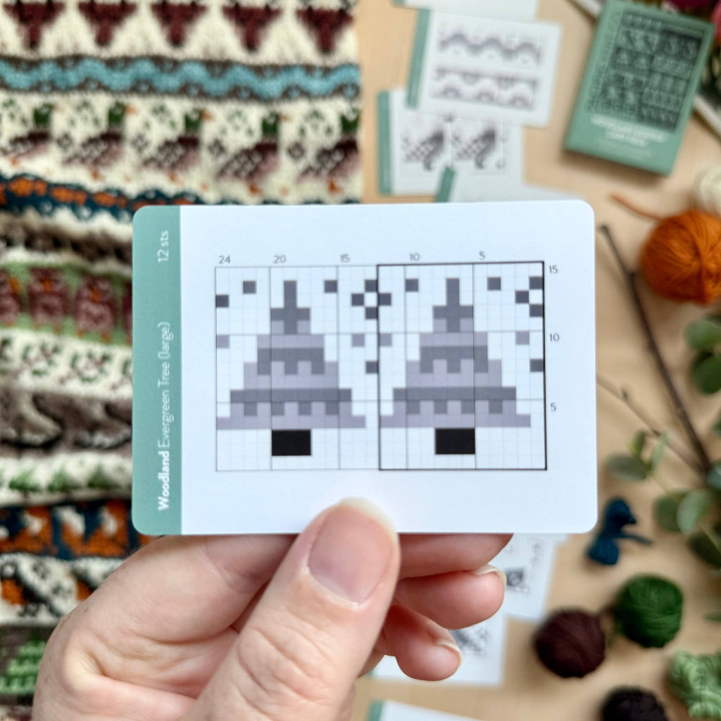 A hand holds a knitting pattern card featuring two gray-and-black knit tree designs, with knitted fabric swatches and yarn balls arranged around it on a tabletop.